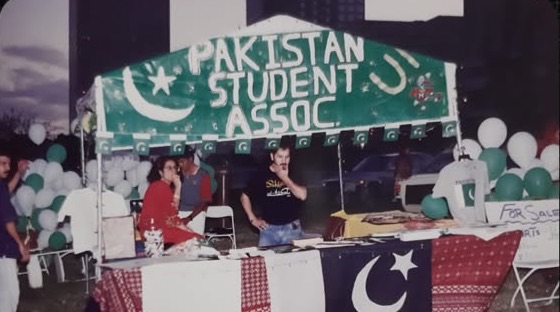 An outdoor PSA stall under a hand-painted green banner reading Pakistan Student Assoc., with green and white balloons and a Pakistani flag on the table